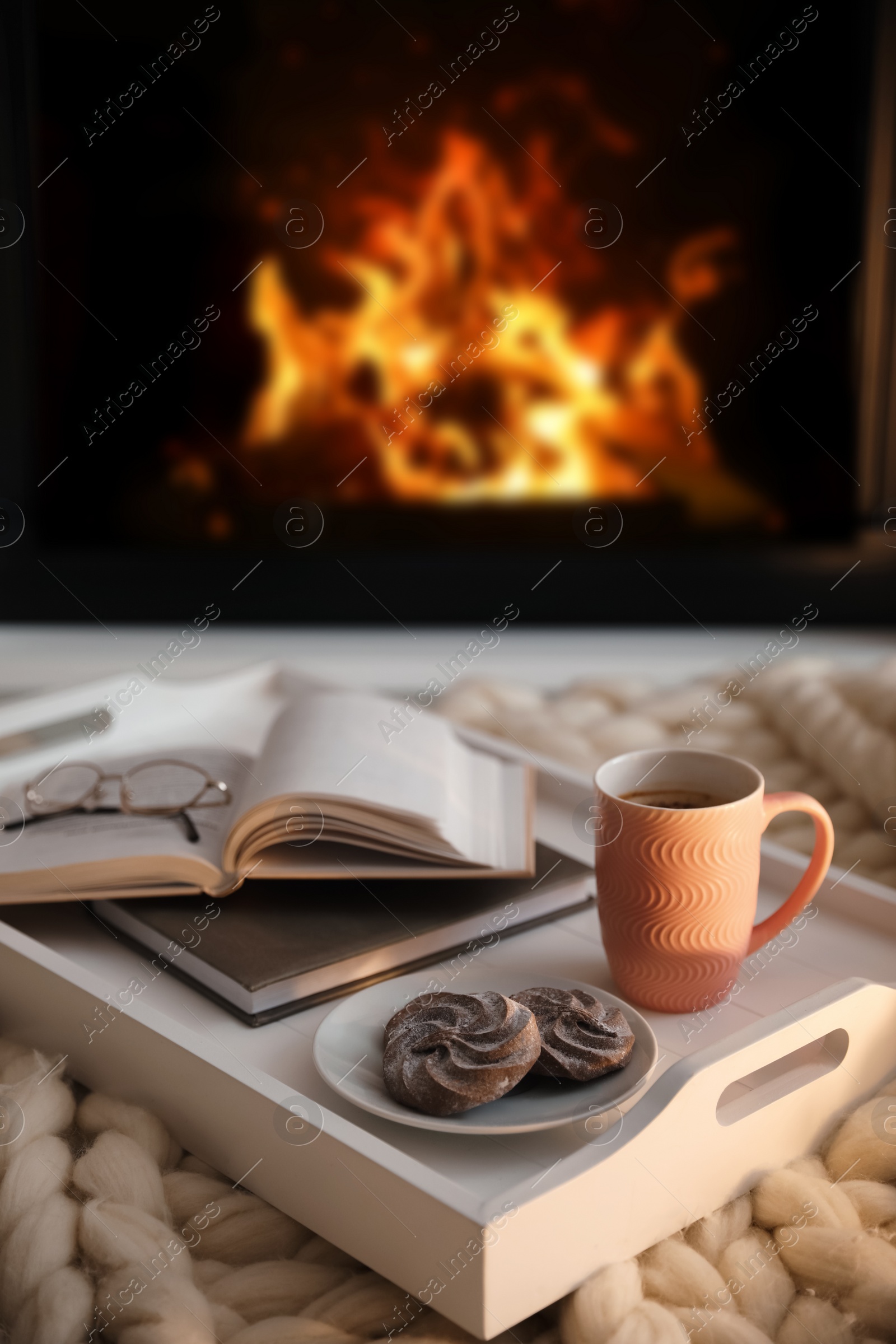 Photo of Cup of coffee, cookies and books on knitted blanket near burning fireplace indoors. Cozy atmosphere