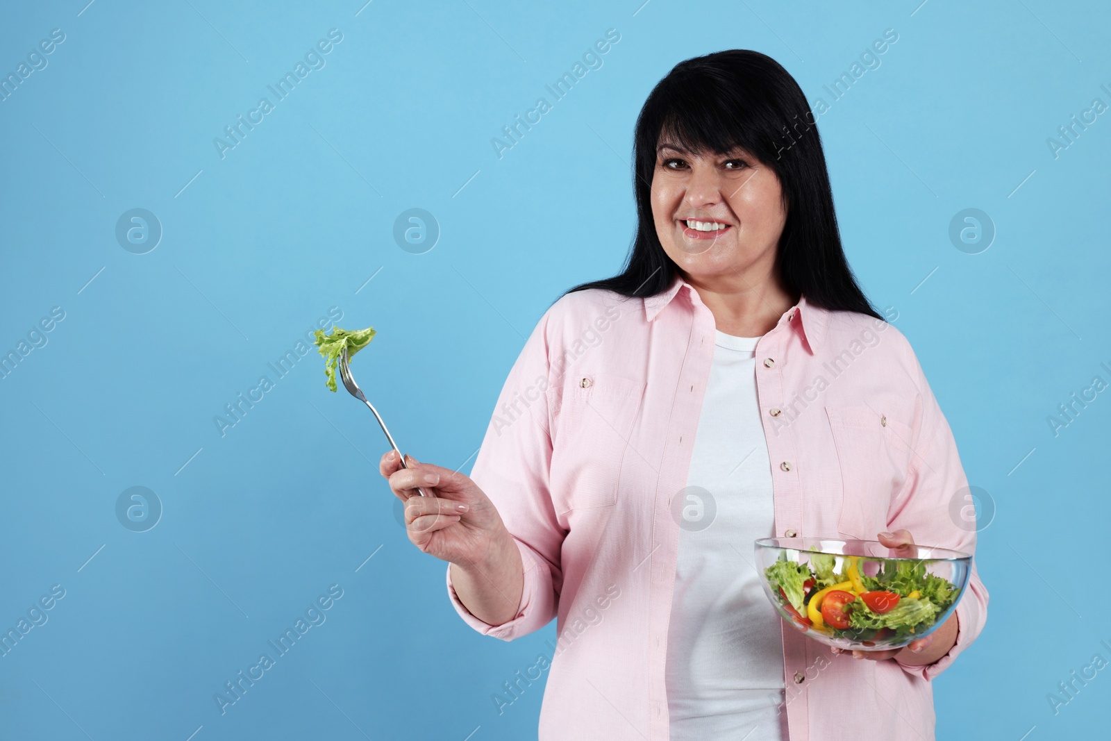 Photo of Beautiful overweight woman eating salad on light blue background, space for text. Healthy diet