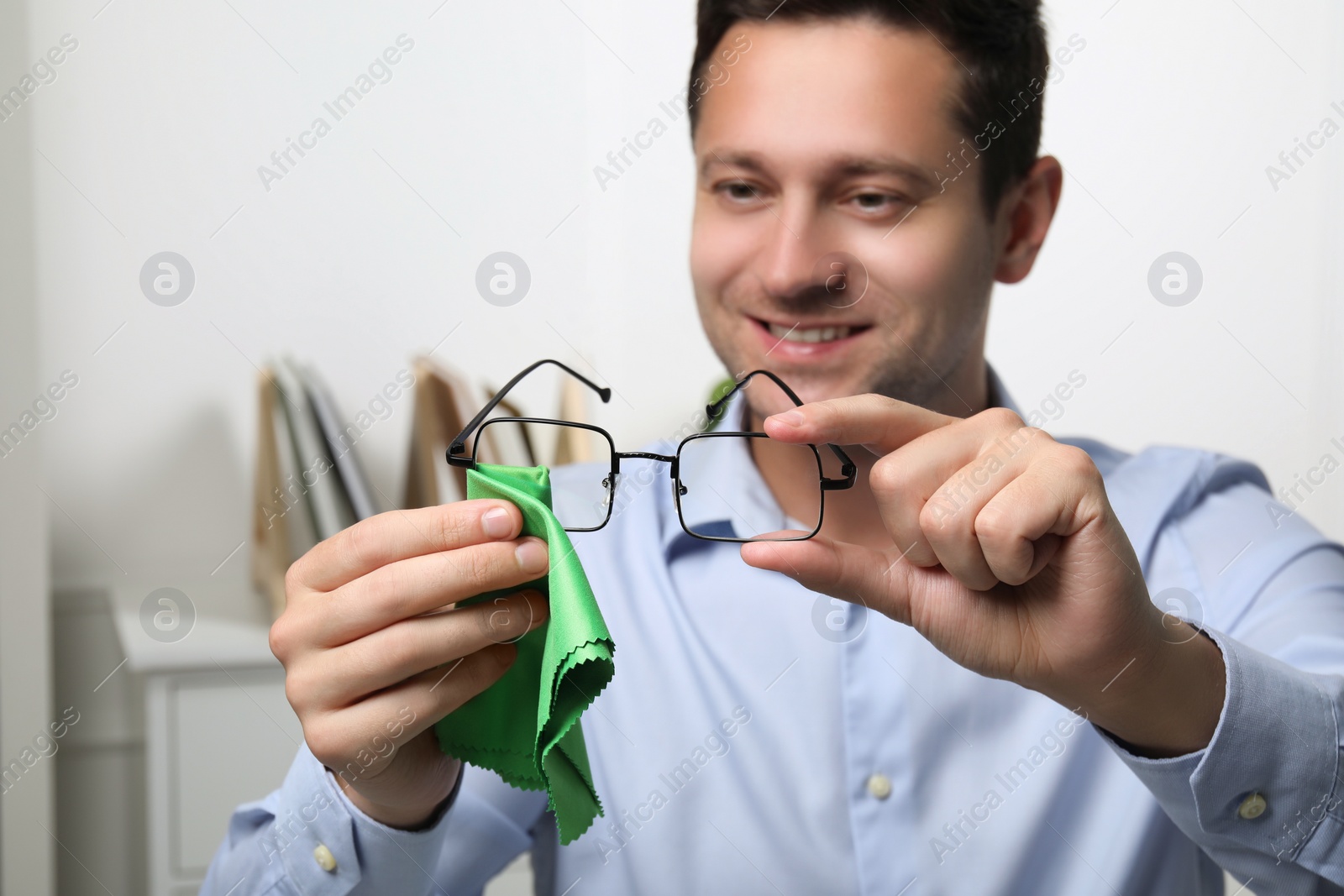 Photo of Happy man wiping glasses with microfiber cloth indoors, selective focus