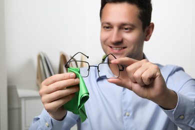 Photo of Happy man wiping glasses with microfiber cloth indoors, selective focus