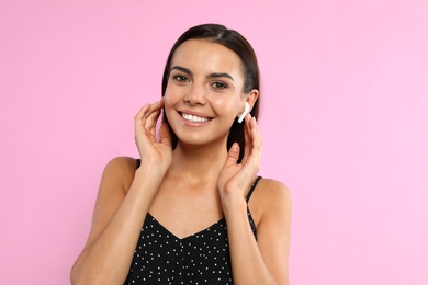 Happy young woman listening to music through wireless earphones on pink background