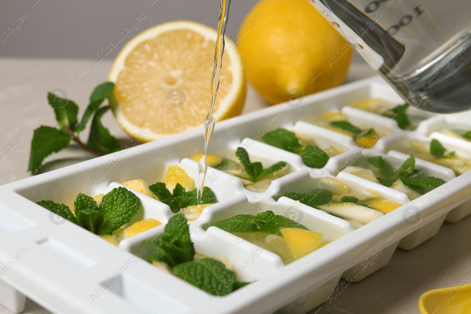 Photo of Pouring water into ice cube tray with mint and lemon on table, closeup