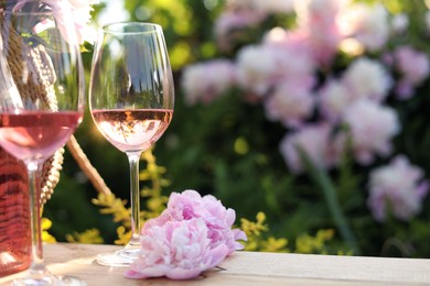 Glasses of rose wine near beautiful peonies on wooden table in garden