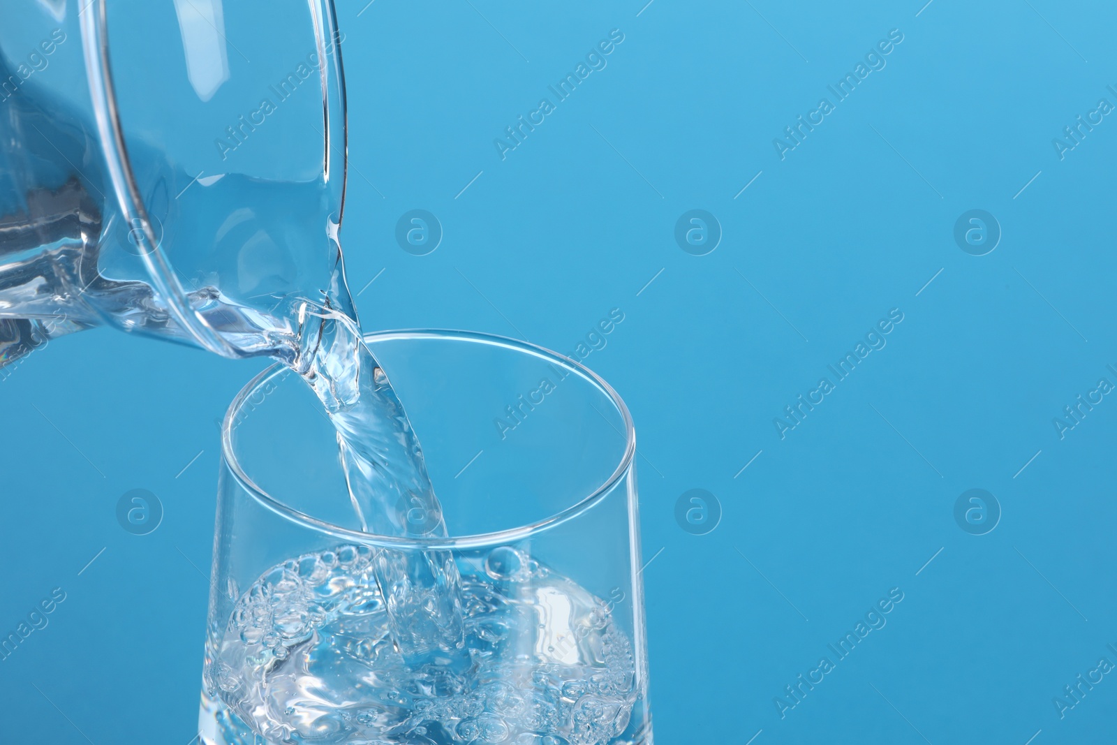 Photo of Pouring water from jug into glass on light blue background, closeup. Space for text