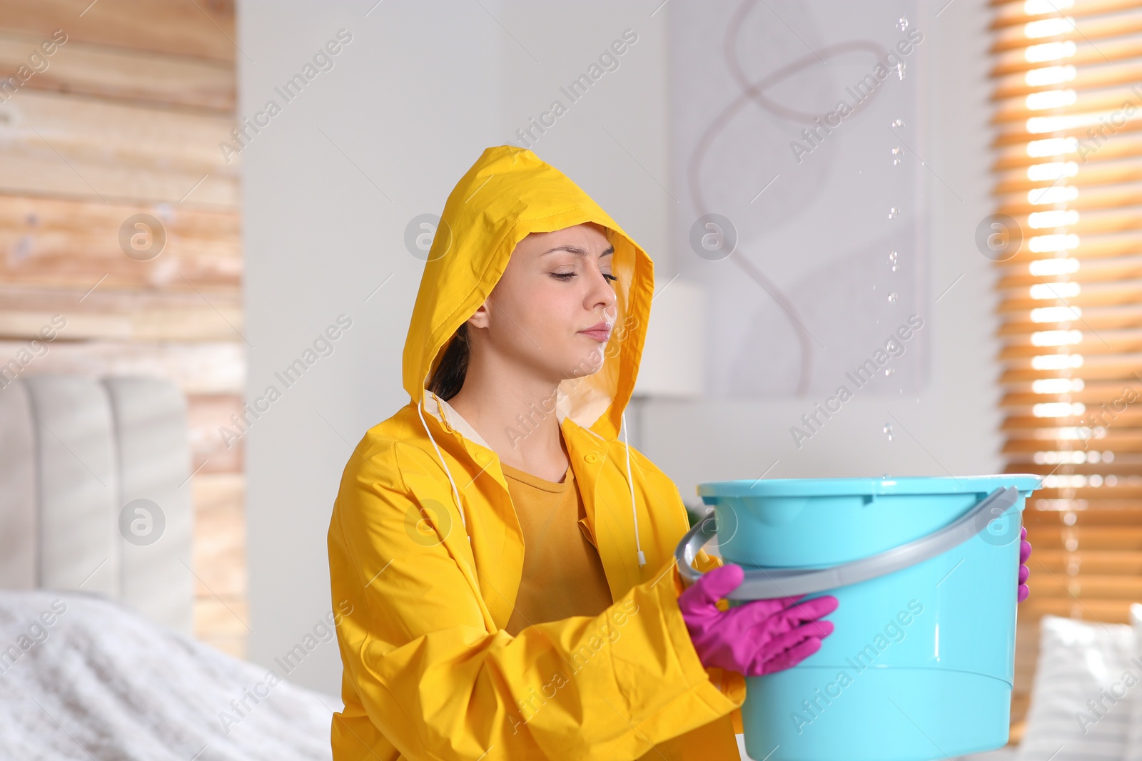 Photo of Young woman in raincoat collecting leaking water from ceiling at home. Time to call roof repair service