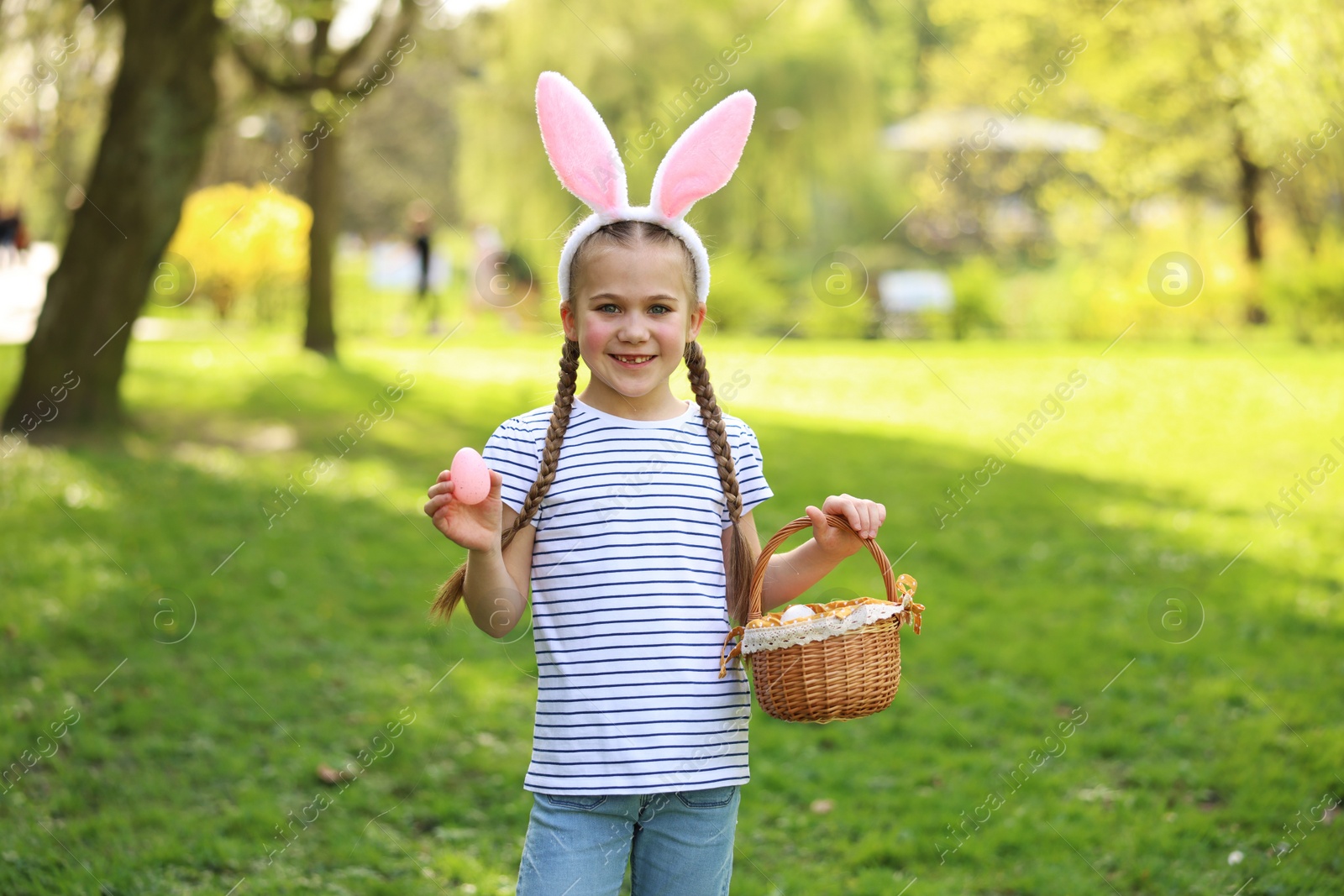 Photo of Easter celebration. Cute little girl in bunny ears holding wicker basket and painted egg outdoors