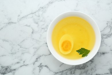 Delicious bouillon with egg and parsley in bowl on white marble table, top view. Space for text