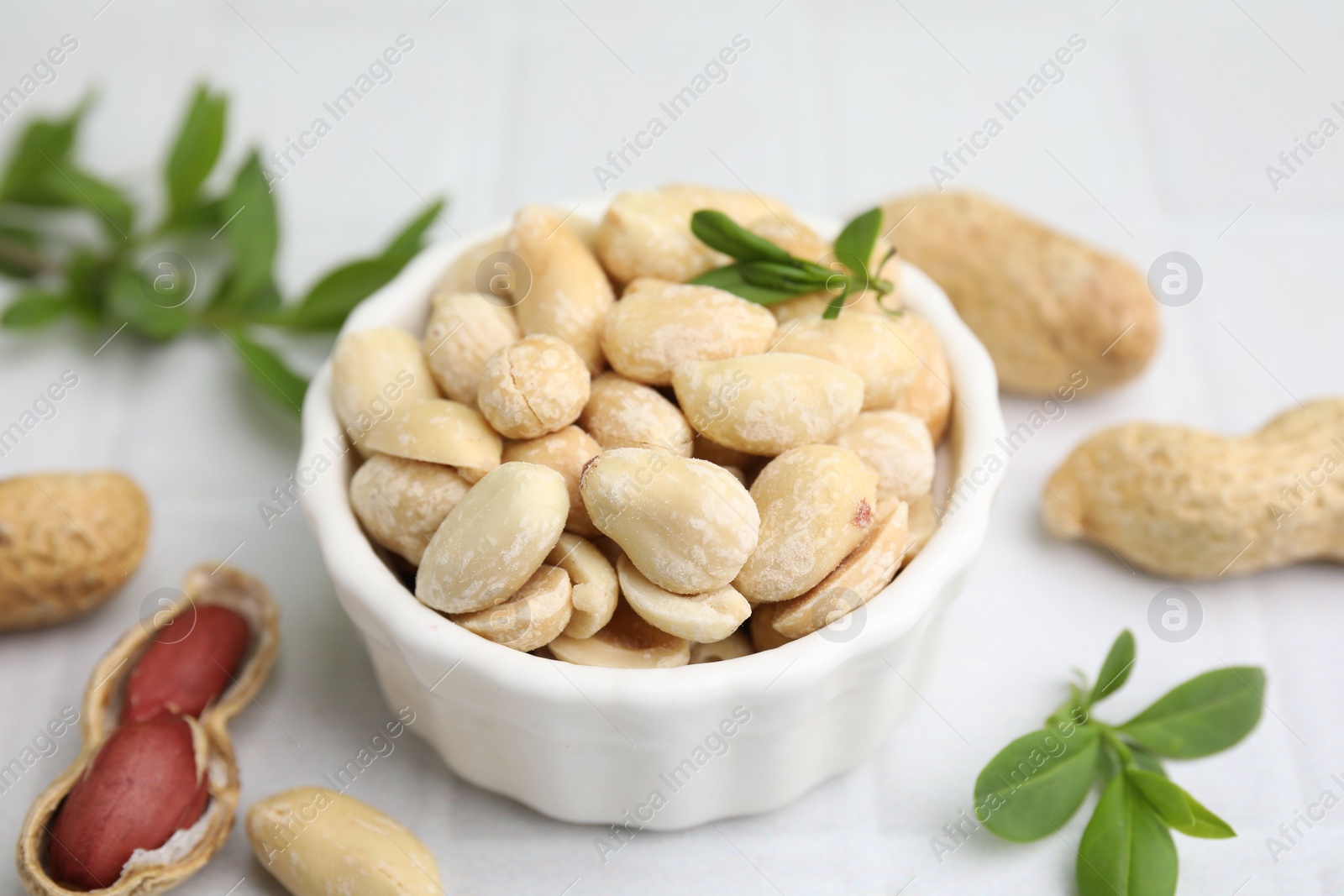 Photo of Fresh peeled peanuts in bowl and leaves on white tiled table, closeup