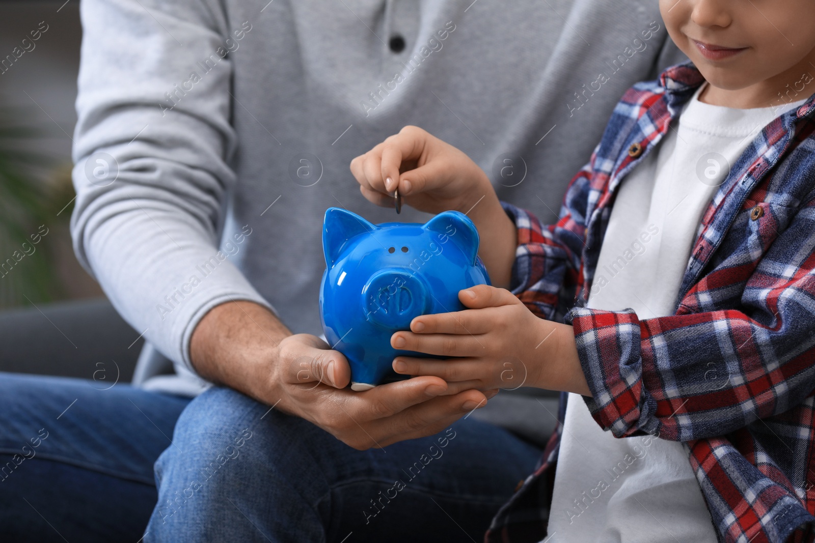 Photo of Little boy with his father putting coin into piggy bank indoors, closeup