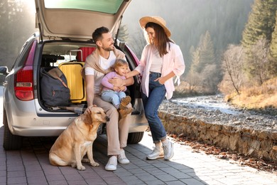 Photo of Parents, their daughter and dog near car outdoors. Family traveling with pet