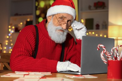 Santa Claus using laptop at his workplace in room decorated for Christmas