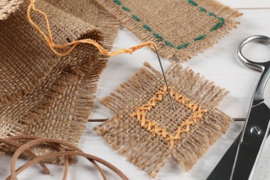 Pieces of burlap fabric with stitches, needle and scissors on white wooden table, closeup