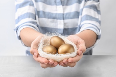 Woman holding nest with golden eggs over table on light background, closeup