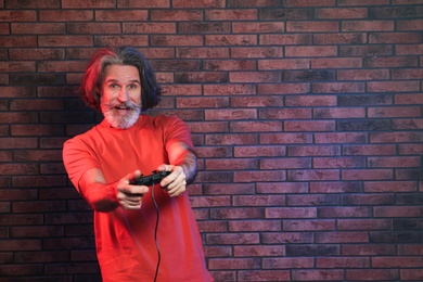 Emotional mature man playing video games with controller near brick wall. Space for text