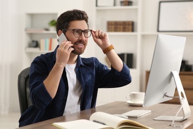 Photo of Home workplace. Happy man talking on smartphone while working with computer at wooden desk in room