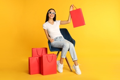 Photo of Beautiful young woman with paper shopping bags in armchair on yellow background