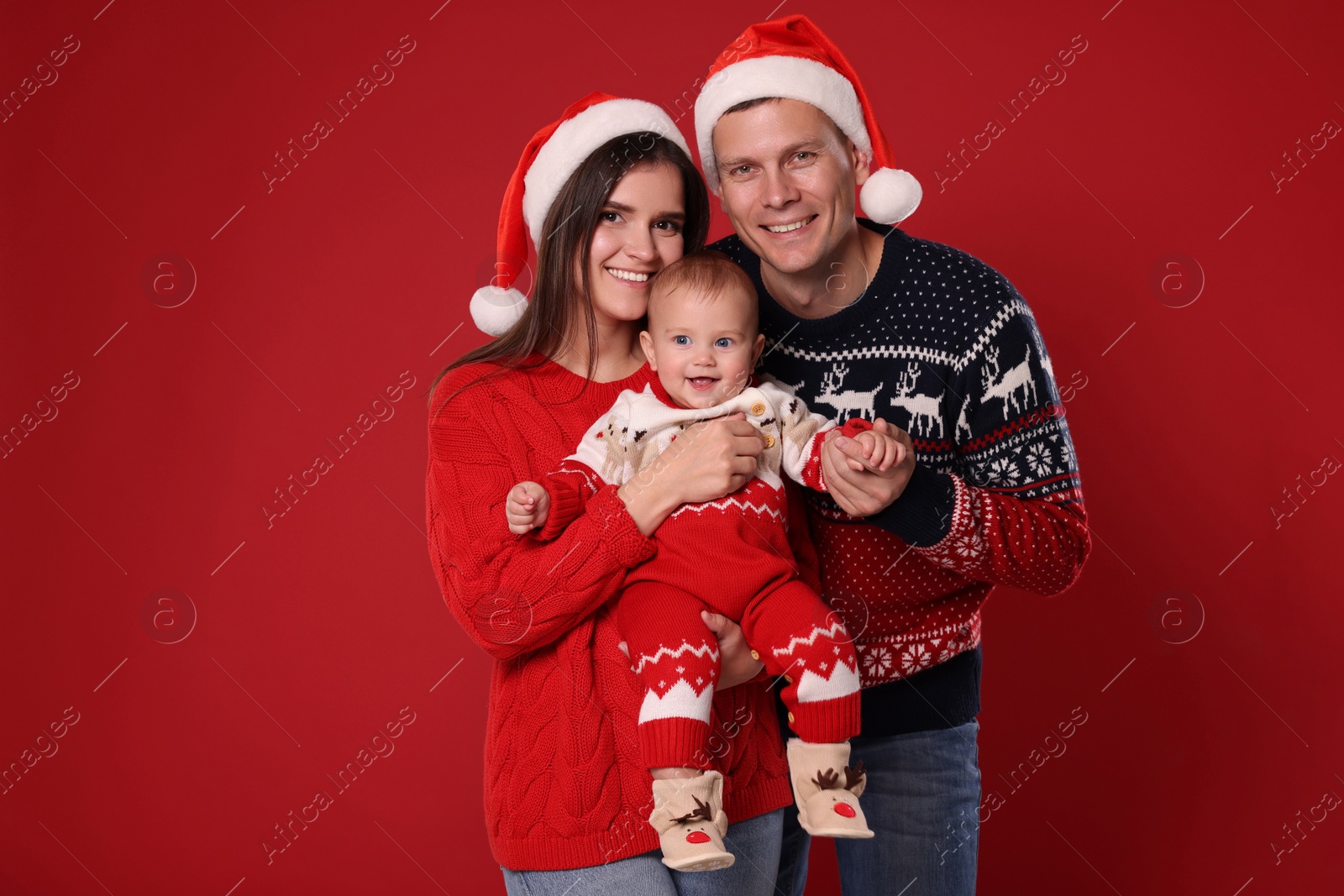 Photo of Happy couple with cute baby in Christmas outfits and Santa hats on red background