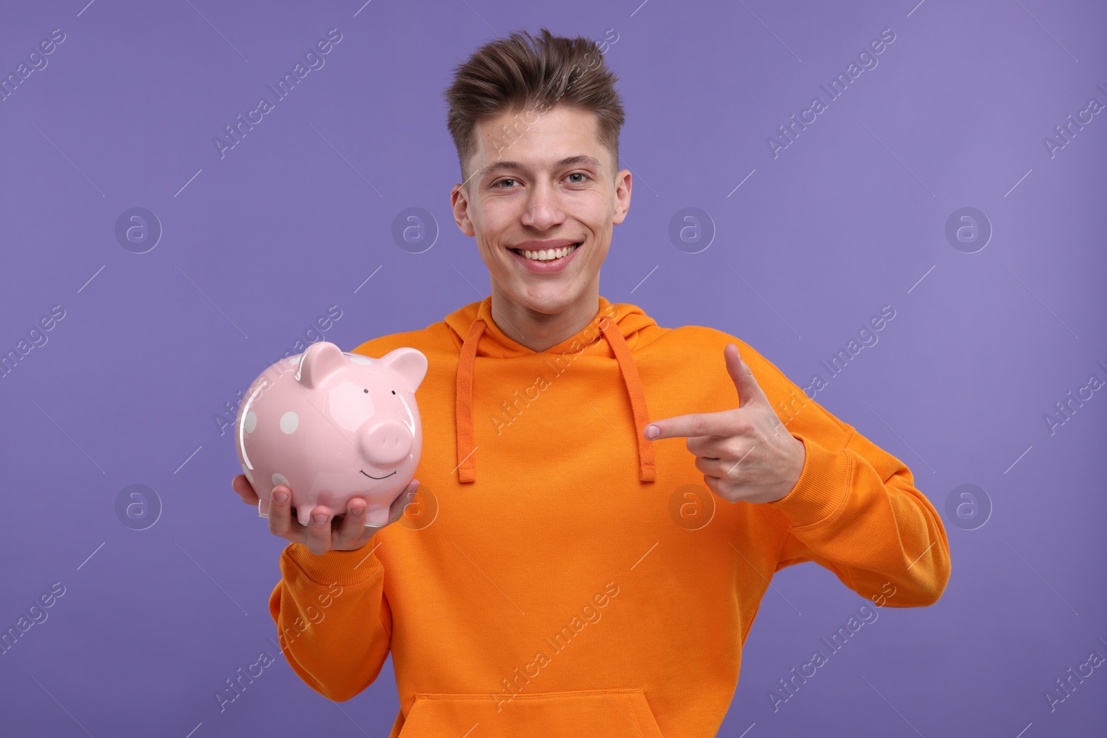 Photo of Happy man pointing at piggy bank on purple background