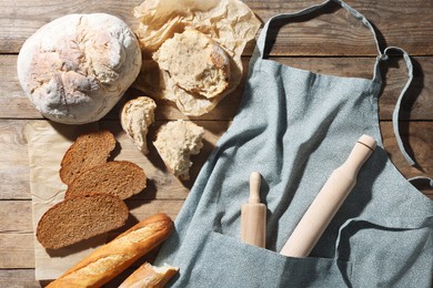 Clean kitchen apron with rolling pins and different types of bread on wooden table, flat lay