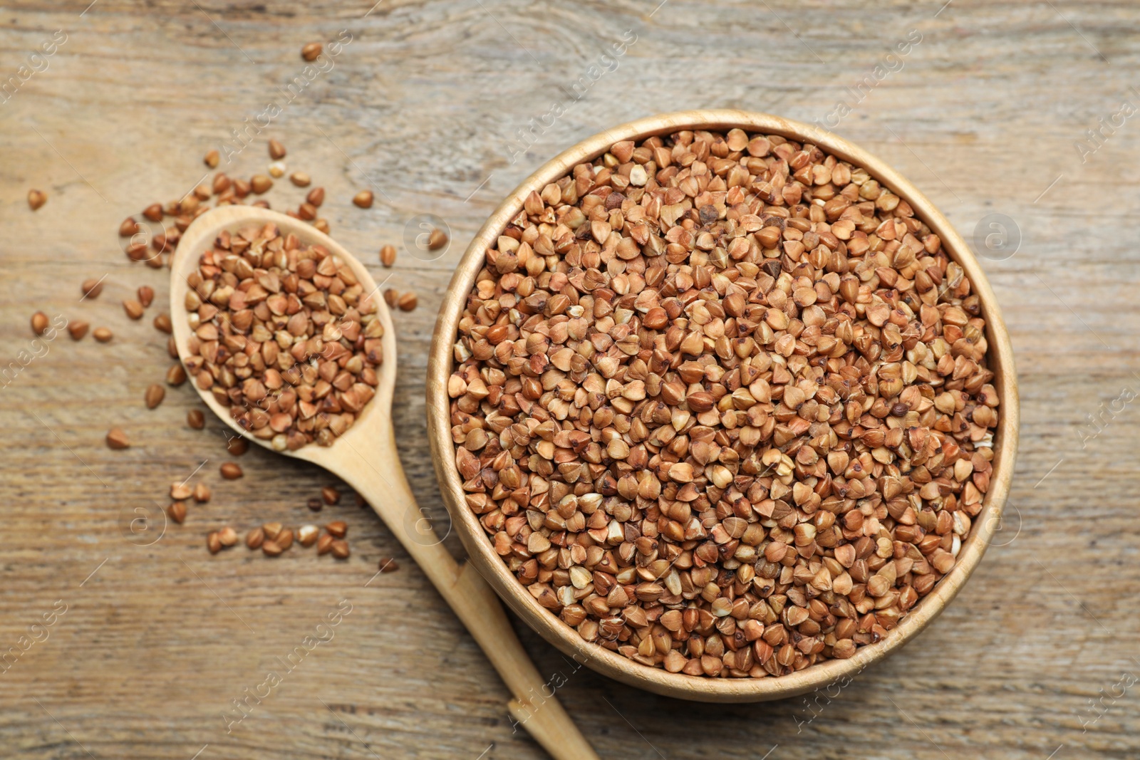 Photo of Bowl and spoon of uncooked buckwheat on wooden table, flat lay