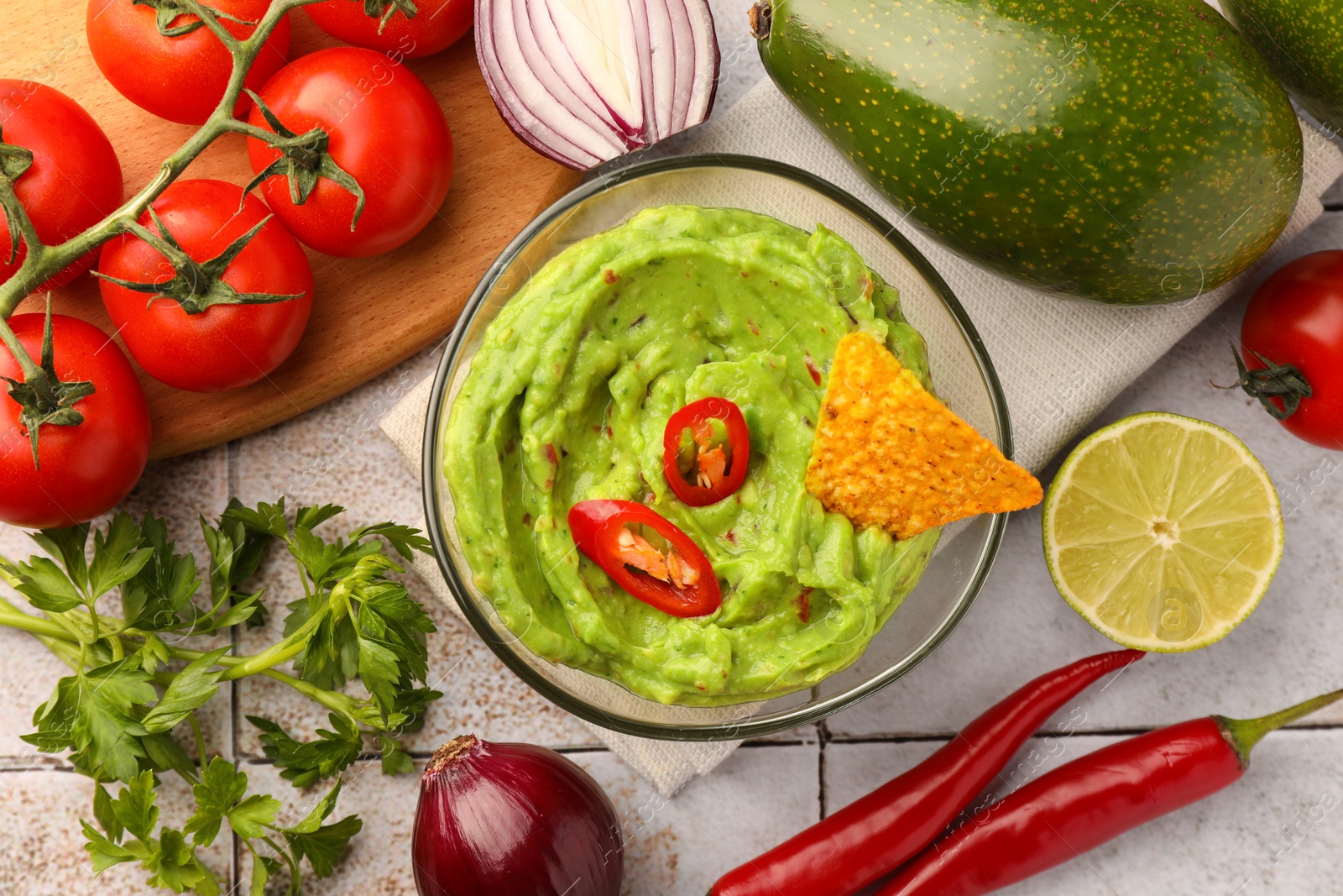 Photo of Bowl of delicious guacamole, nachos chip and ingredients on white tiled table, flat lay