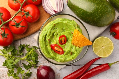 Bowl of delicious guacamole, nachos chip and ingredients on white tiled table, flat lay