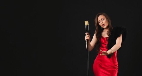 Beautiful young woman in stylish dress with microphone singing on black background