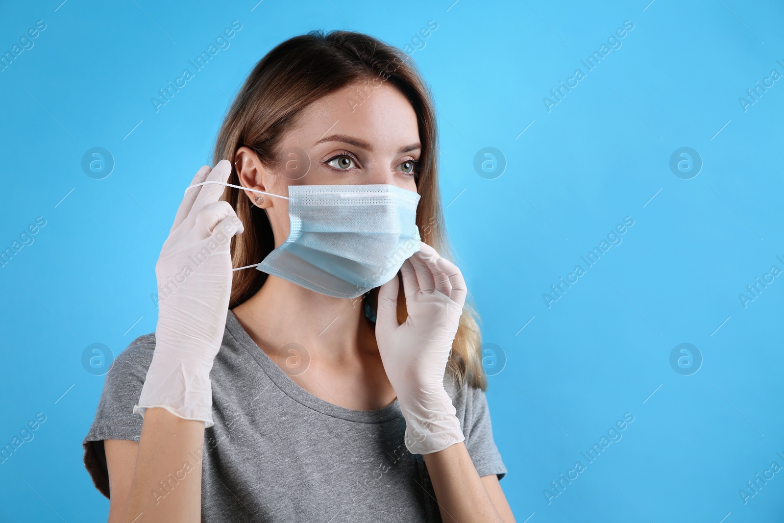 Photo of Woman in medical gloves putting on protective face mask against light blue background