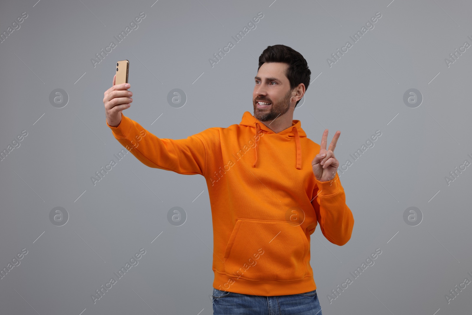 Photo of Smiling man taking selfie with smartphone and showing peace sign on grey background