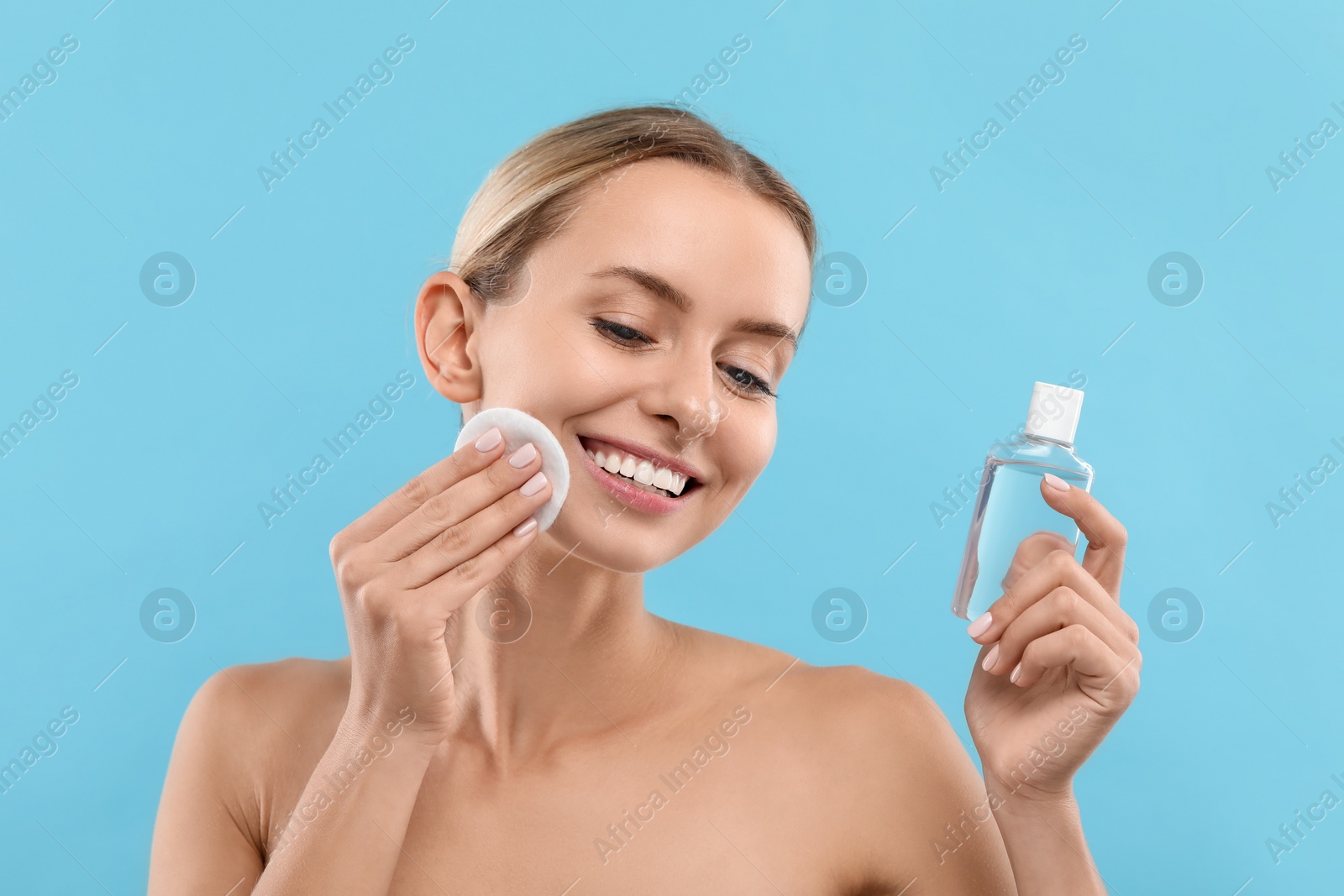 Photo of Smiling woman removing makeup with cotton pad and holding bottle on light blue background