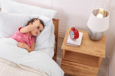 Little girl lying in bed and looking at alarm clock on bedside table indoors, above view