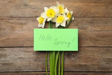 Photo of Green card with words HELLO SPRING and narcissus on wooden table, flat lay