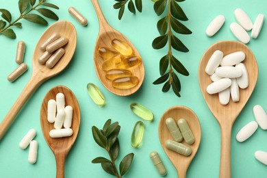 Different vitamin capsules in spoons and leaves on turquoise background, flat lay