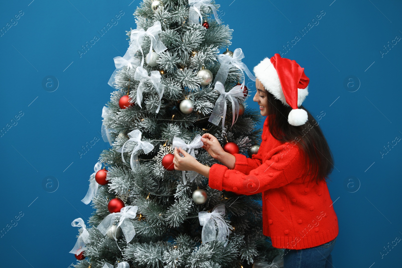 Image of Beautiful young woman in Santa hat decorating Christmas tree on blue background