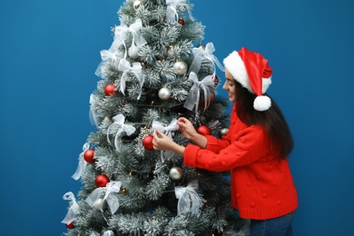 Beautiful young woman in Santa hat decorating Christmas tree on blue background