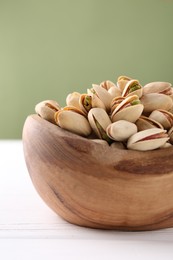 Photo of Tasty pistachios in bowl on white wooden table against olive background, closeup