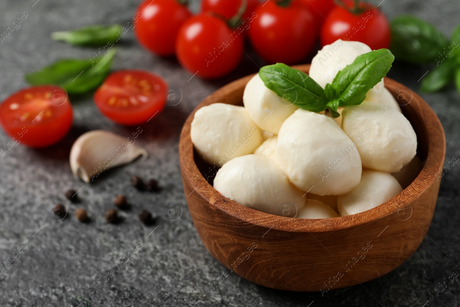 Photo of Delicious mozzarella balls in wooden bowl with basil leaves and tomatoes on light gray table, closeup. Space for text