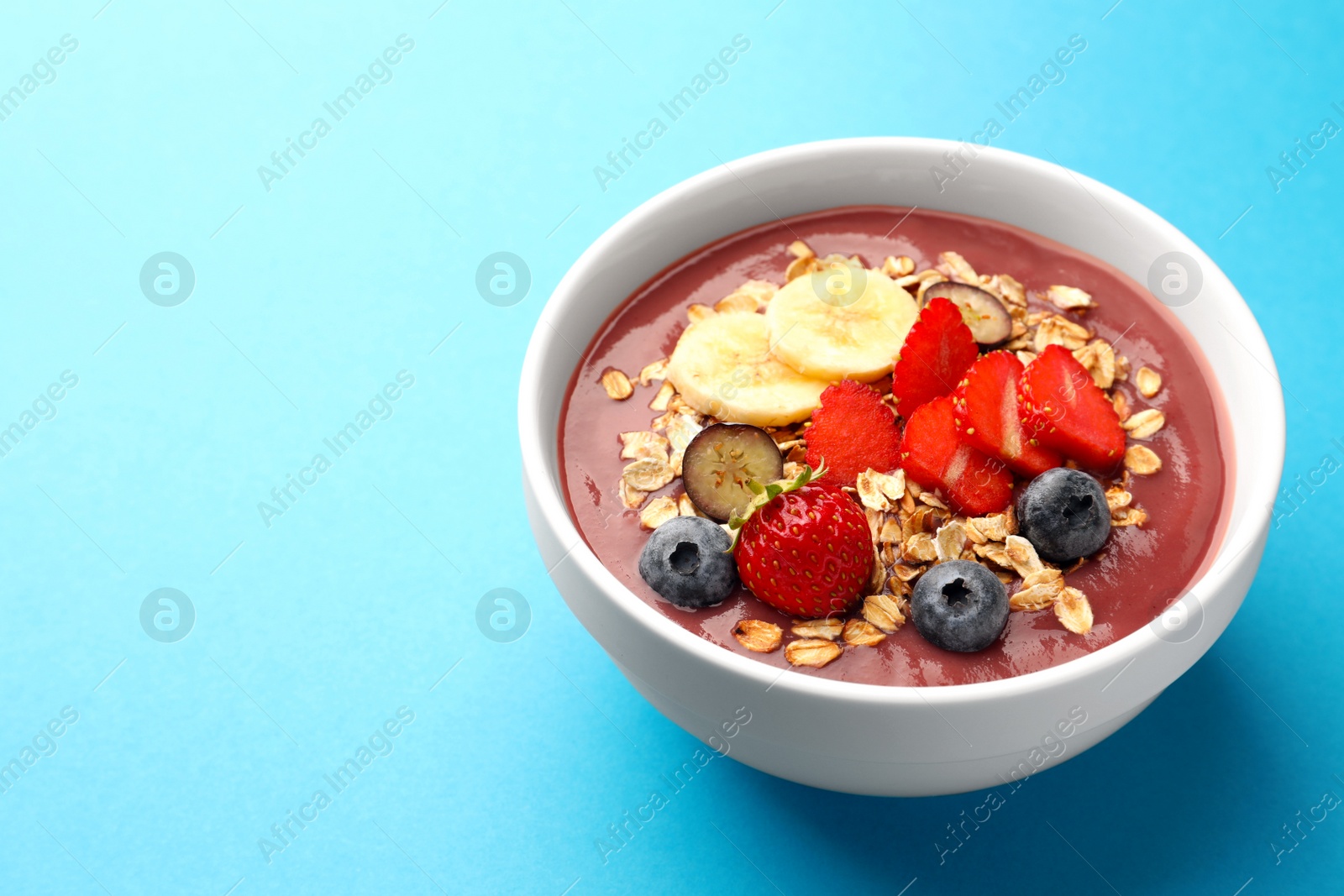 Photo of Delicious smoothie bowl with fresh berries, banana and granola on light blue background. Space for text
