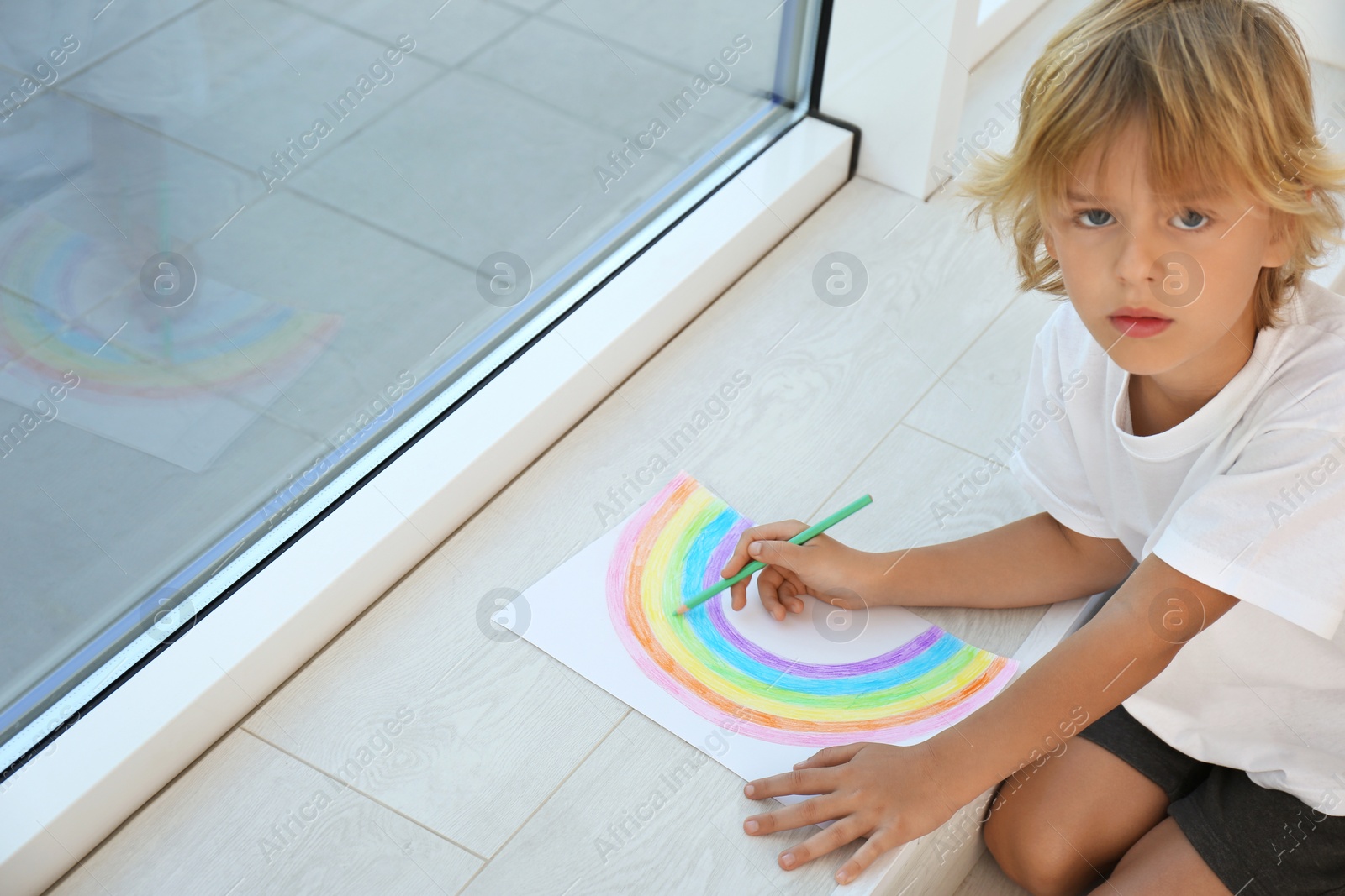 Photo of Little boy drawing rainbow near window indoors. Stay at home concept