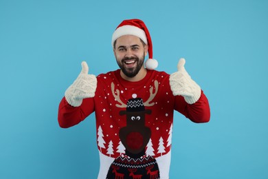 Photo of Happy young man in Christmas sweater, Santa hat and knitted mittens showing thumbs up on light blue background