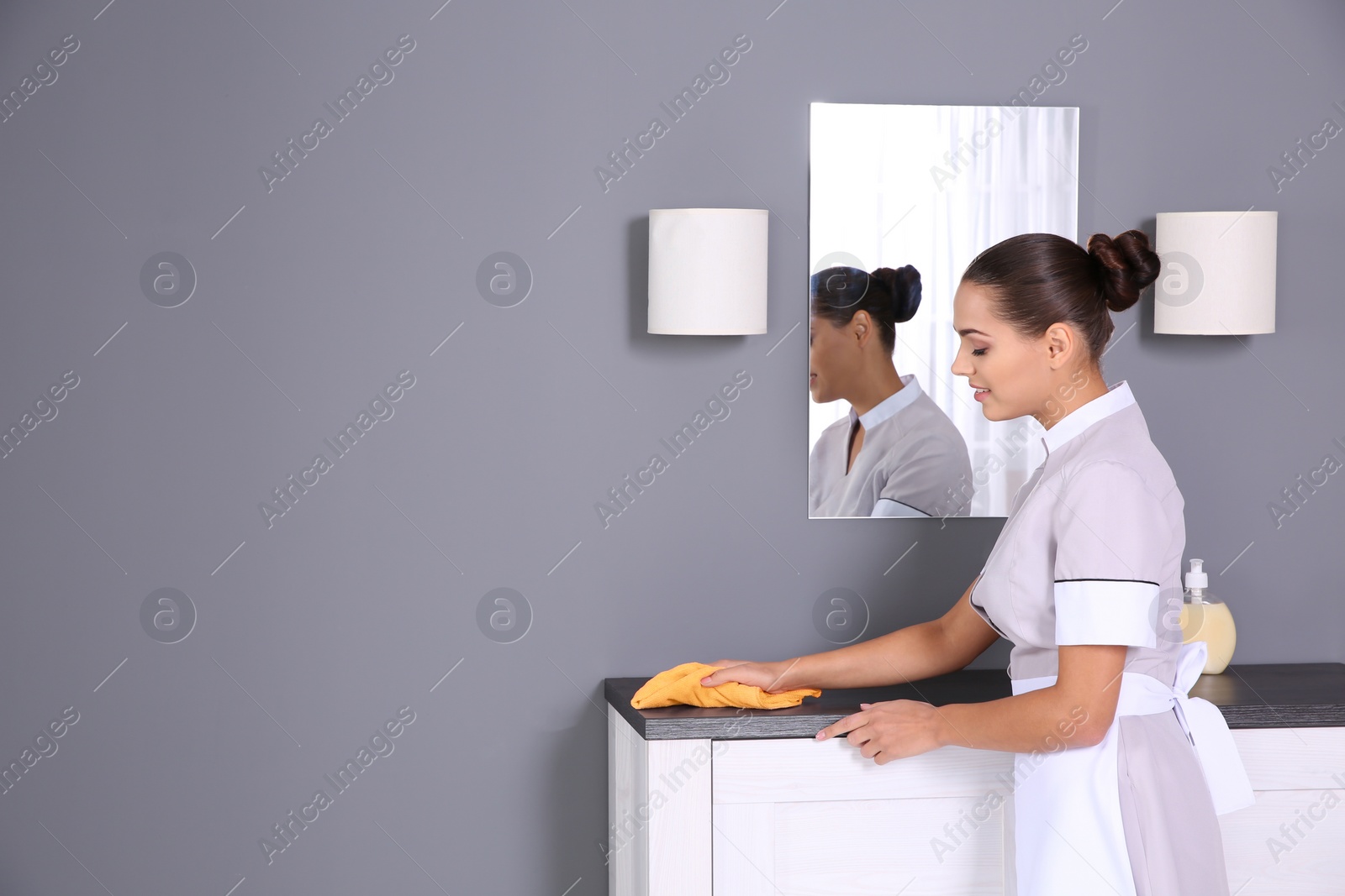 Photo of Young chambermaid wiping dust from furniture with rag in bathroom. Space for text