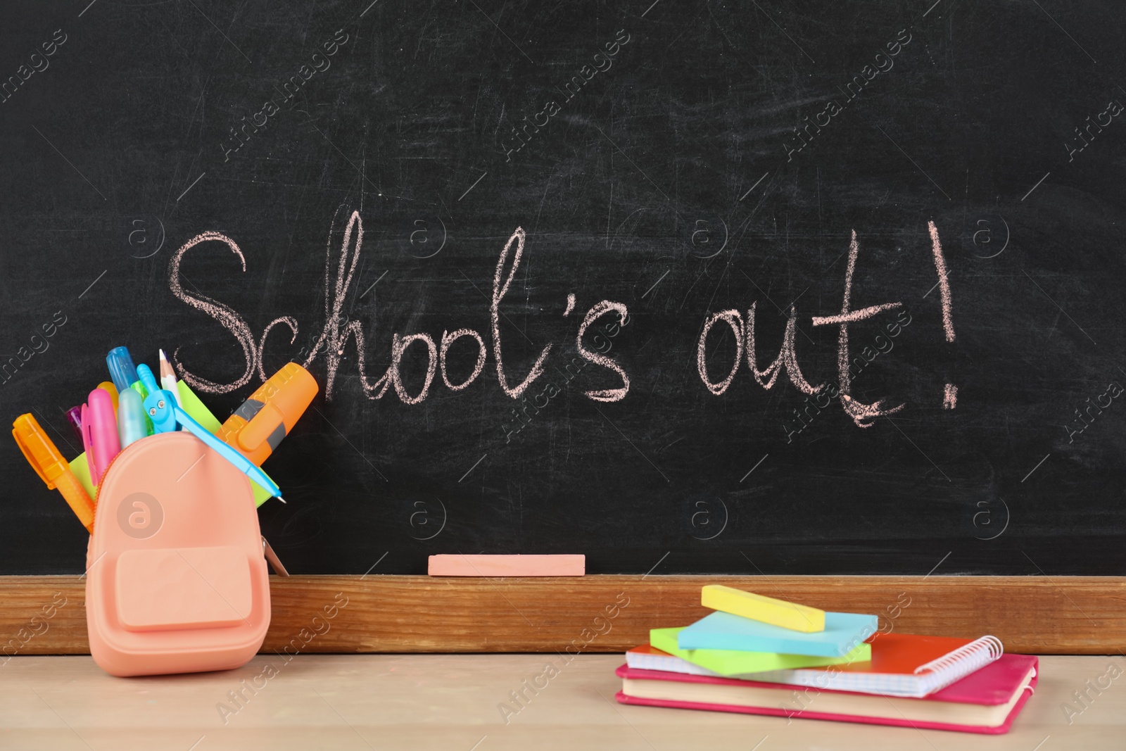 Photo of Pieces of color chalk and stationery on table near blackboard with text School's Out. Summer holidays