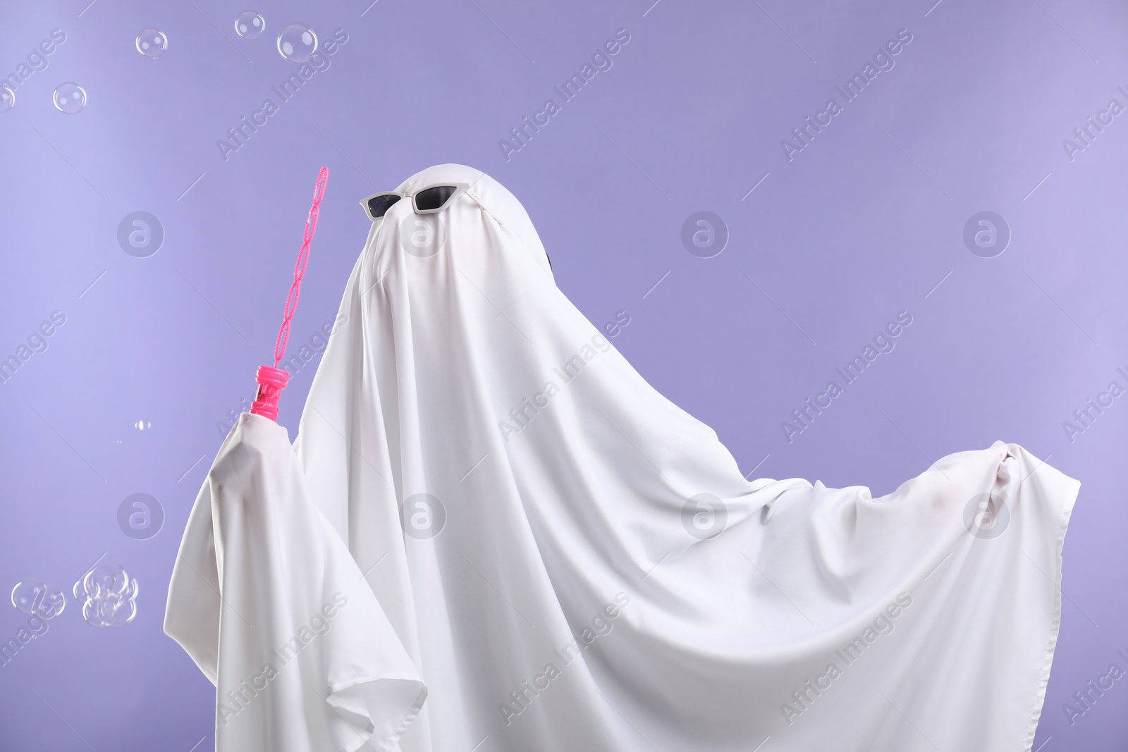 Photo of Funny ghost. Person in white sheet and sunglasses blowing soap bubbles on violet background