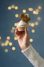 Photo of Woman holding tasty Christmas cupcake with gingerbread man cookie against blurred festive lights, closeup