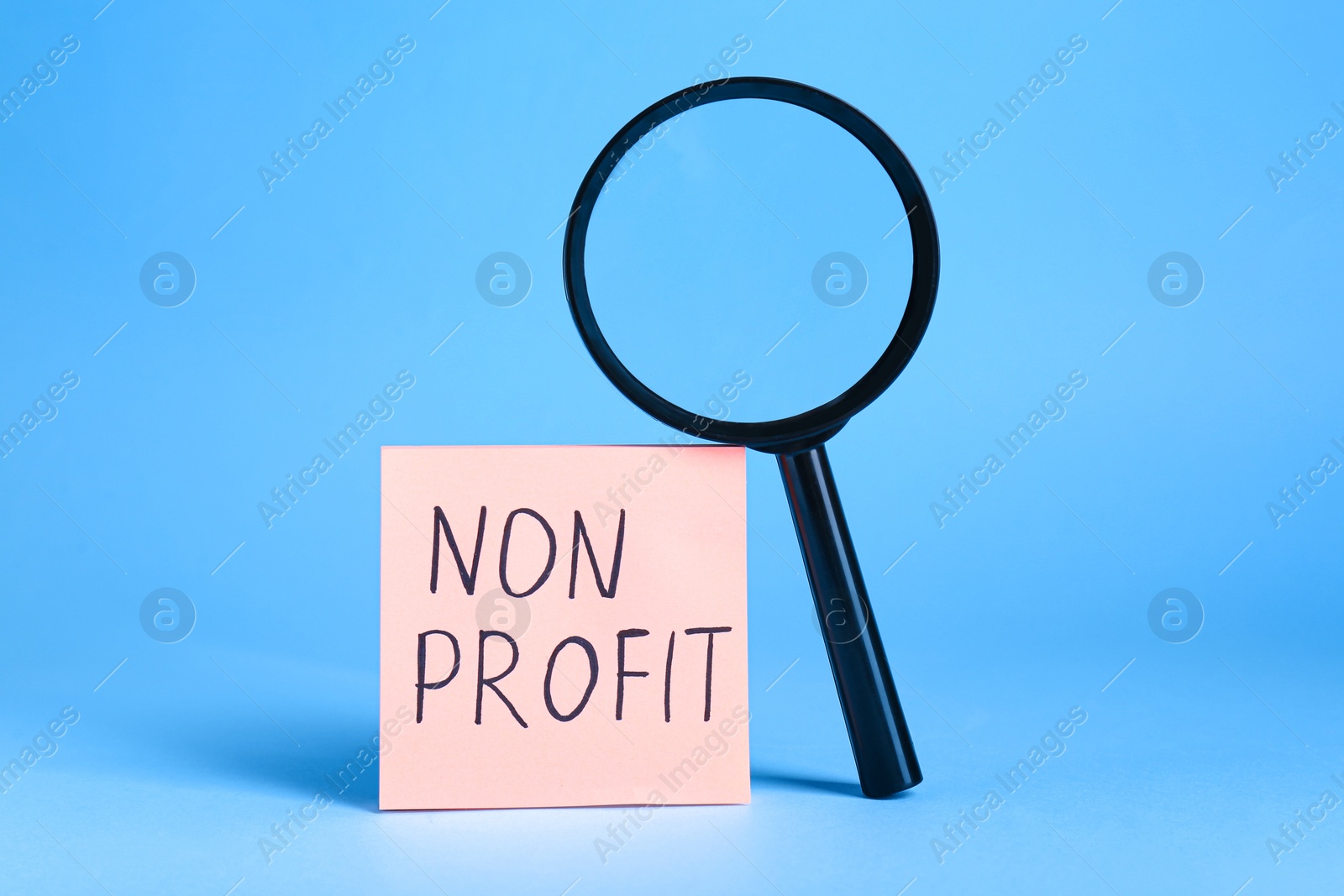 Photo of Paper note with phrase Non Profit and magnifying glass on light blue background