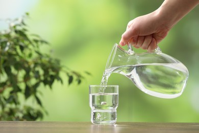 Photo of Woman pouring fresh water from jug into glass at wooden table against blurred green background, closeup. Space for text