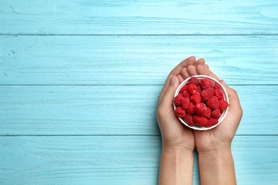 Photo of Woman holding bowl with delicious ripe raspberries at blue wooden table, top view. Space for text