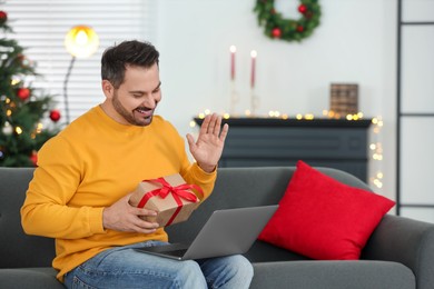 Photo of Celebrating Christmas online with exchanged by mail presents. Man with gift waving hello during video call on laptop at home
