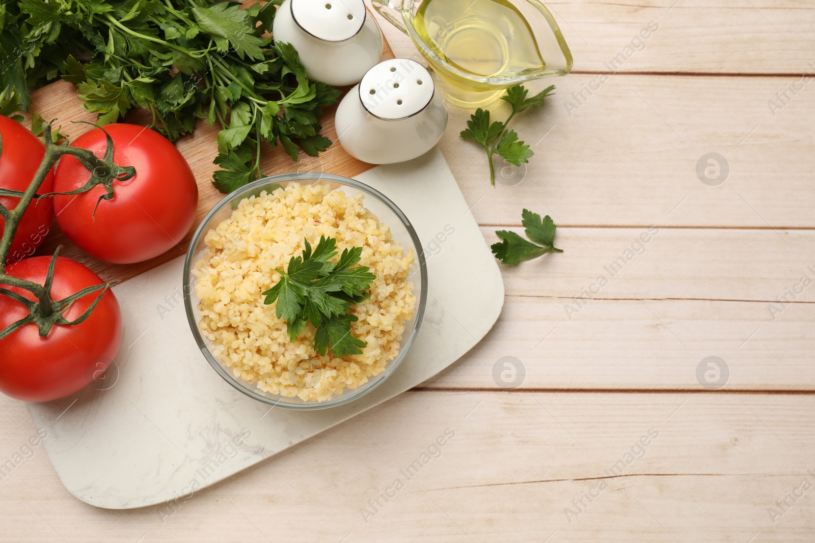 Photo of Delicious bulgur with parsley in bowl, tomatoes, oil and spices on wooden table, top view. Space for text