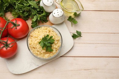 Delicious bulgur with parsley in bowl, tomatoes, oil and spices on wooden table, top view. Space for text